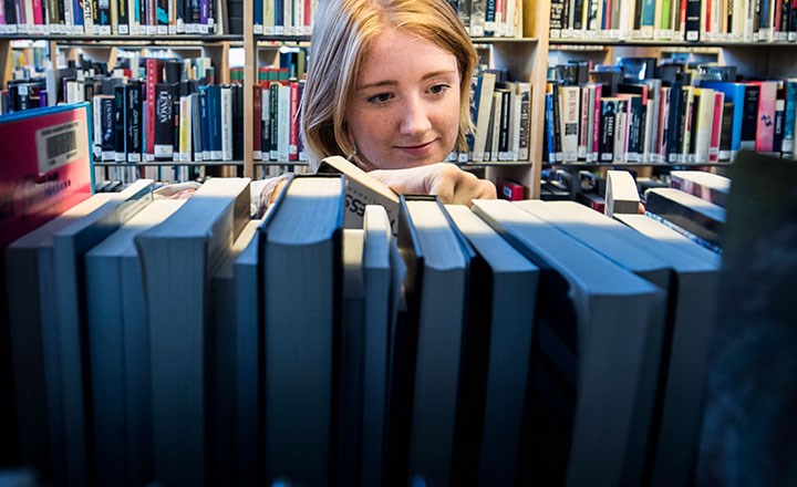 En kvinna står mellan bokhyllor i ett bibliotek. Hennes ansikte syns mellan bokhyllorna. 