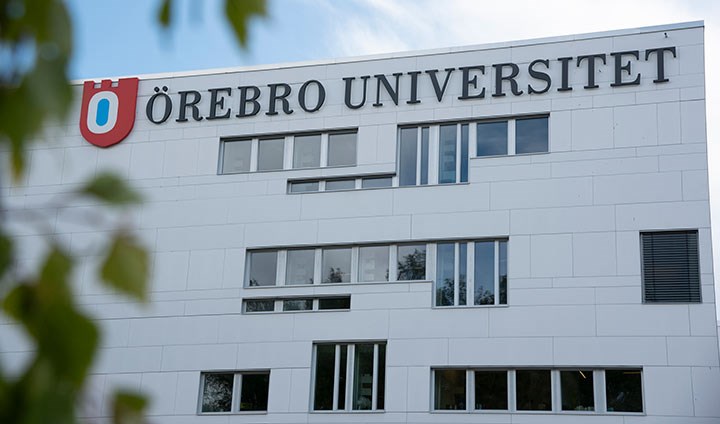 Det vita Novahuset med en stor skylt där det står "Örebro universitet". I förgrunden syns suddiga gröna löv på ett träd.