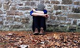 A young person hunched up and holding their head between arms, sitting on leaves on the ground.