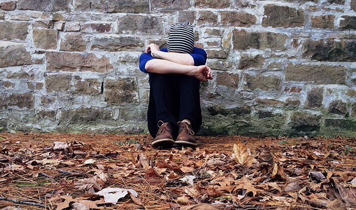 A young person hunched up and holding their head between arms, sitting on leaves on the ground.