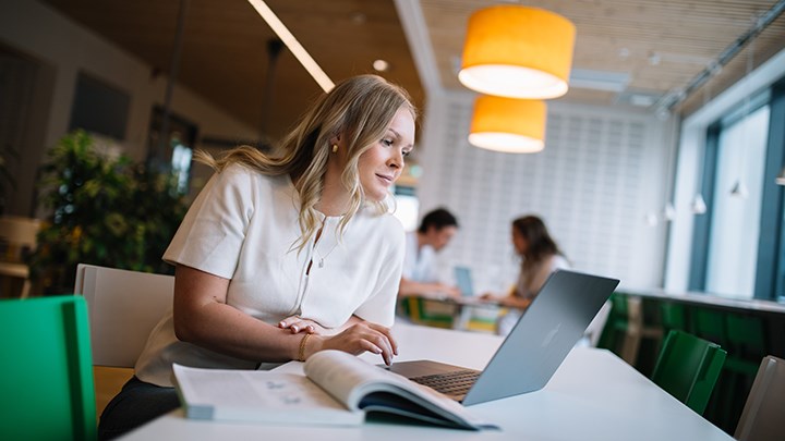 En kvinna sitter vid ett bord med en laptop där skärmen är öppen. En uppslagen bok finns på bordet. 