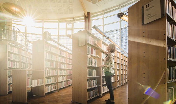 Student sträcker sig efter en bok i ett ljust bibliotek med många bokhyllor.