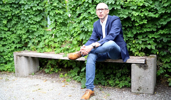 Magnus Hansson sitting outside on a bench with his legs crossed, in front of a wall of plants.