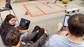 Three persons using computers and sitting in front of mini-robots.