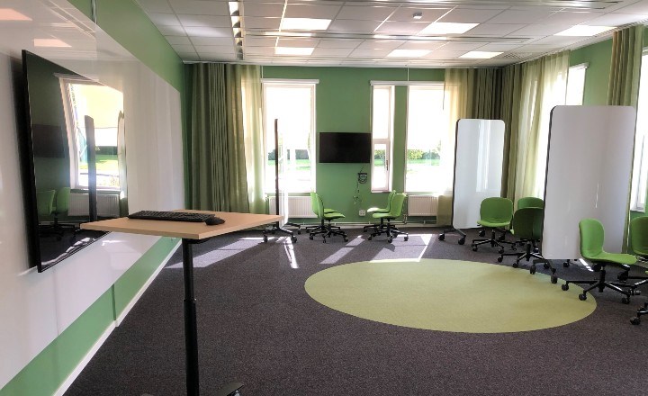 Rum med gröna väggar, whiteboard och Tv-skärm, ett ståbord och en heltäckningsmatta med grön prick i mitten