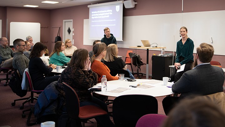 Elin Vimefall sitter längst fram vid en whiteboard. En grupp med åhörare sitter vid runda bord och lyssnar på henne.