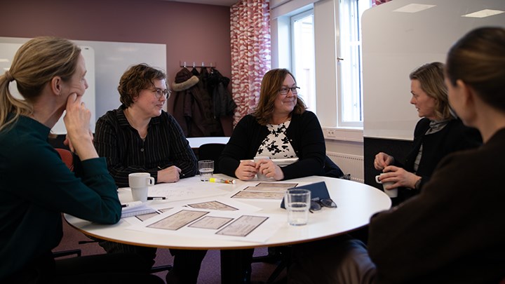 Fem kvinnor sitter runt ett runt bord och samtalar.