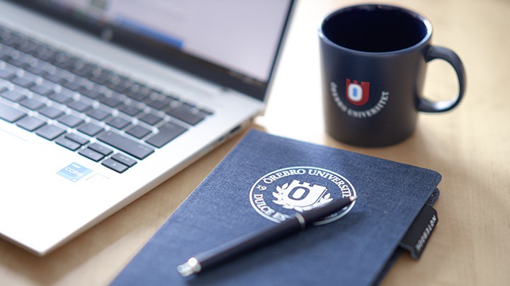 A computer, a dark blue mug with the university logo, and a dark blue notebook with the university logo and a pen on it.
