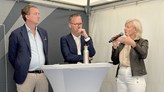 Två män och en kvinna samtalar. De står vid ett vitt ståbord. Kvinnan har en mikrofon i handen. 