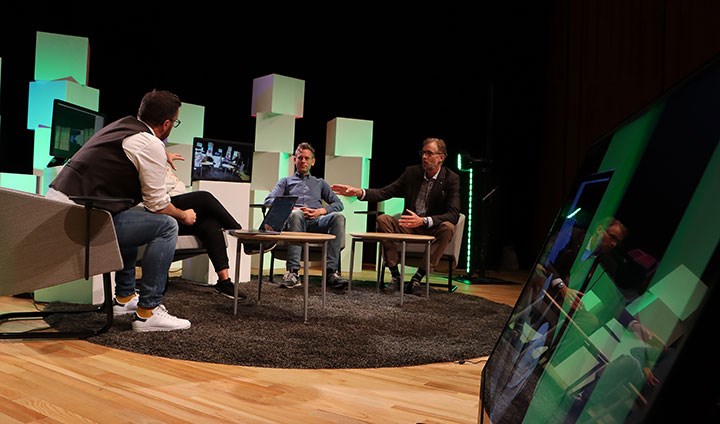 Fyra personer sitter runt ett bord på en scen och pratar. I förgrunden syns en tv-skärm.