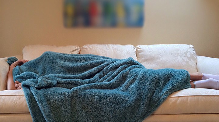  A young person lies on a sofa with a blanket over his head.