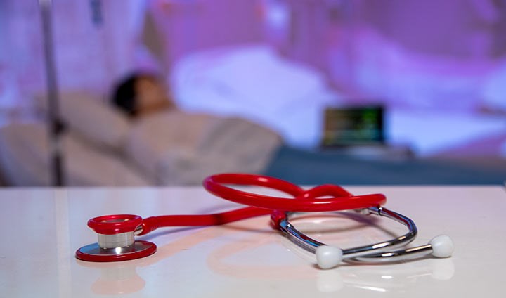 Ett rött stetoskop i förgrunden, i bakgrunden en blå sjukhusmiljö med en suddig säng.