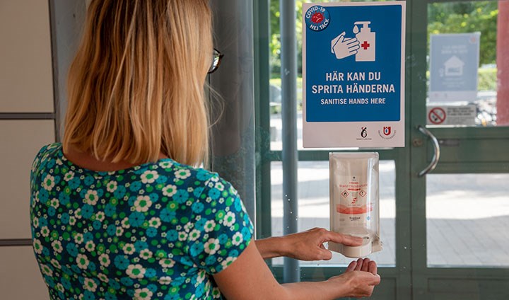 En kvinna tar handsprit ur en automat fäst vid en entré. På en skylt står det "Här kan du sprita händerna".