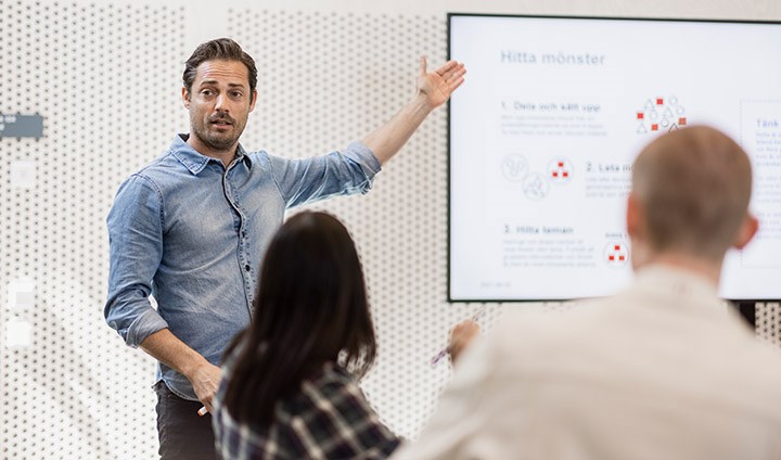 En man föreläser för en grupp studenter. Han pekar på en powerpoint-presentation på en tv-skärm.