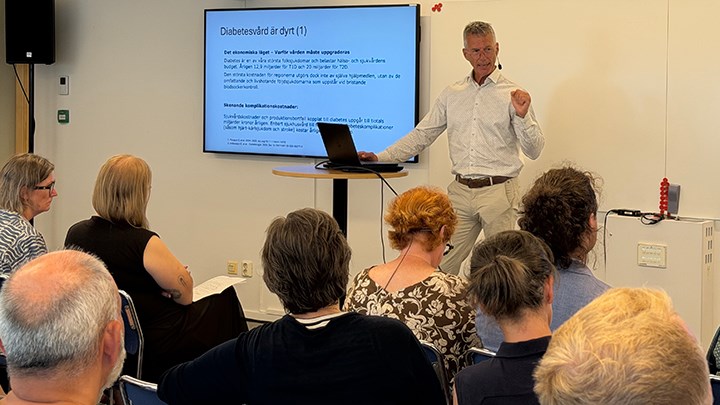 Johan Jendle står vid en storbildsskärm. Han har en vit skjorta och beige byxor.
