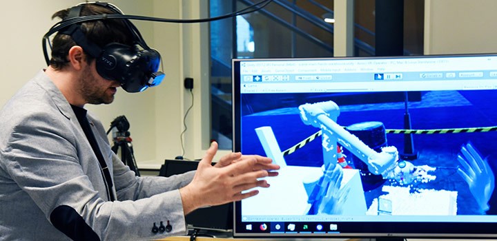 Foto på person som står med VR-glasögon samt en skärm i bakgrunden som visar en robot.