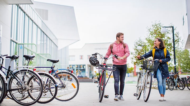 Två studenter går på campus med varsin cykel och pratar med varandra.