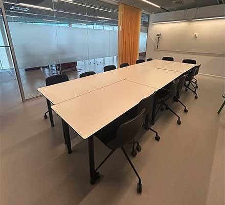 Vy 2 - Rektangulära bord med stolar runt framför en whiteboardtavla. Glasvägg vid sidan.