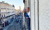 Två kvinnor tittar ut från en fransk balkong. I bakgrunden syns en trafikerad gata.