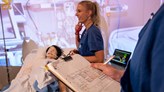 Foto på en person i sjuksköterskekläder står vid en patientsäng där det ligger en docka.