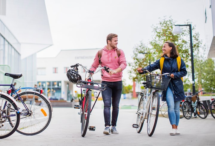 En kvinnlig och en manlig student som går och leder sina cyklar utanför Nova huset 