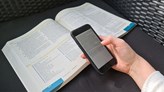 Foto på en uppslagen bok och en hand som håller i en mobiltelefon som visar en e-bok.