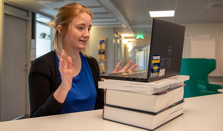 A girl sitting in a virtual meeting. She has put some thick books under her laptop.