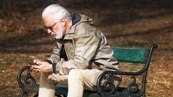 En äldre man sitter på en parkbänk med en mobiltelefon i handen. 