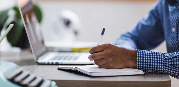 Person sitting with a computer and writing on a notebook.