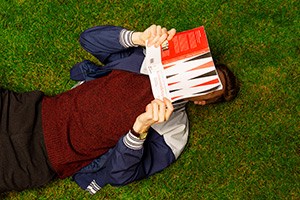 Person som ligger ner i gräset och håller i en bok som täcker ansiktet