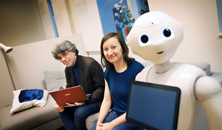 En man, en kvinna och en robot sitter bredvid varandra i en soffa.
