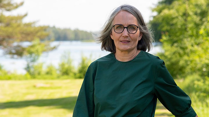 Åsa Öström står i naturen med gräsmatta, träd och vatten i bakgrunden. 
