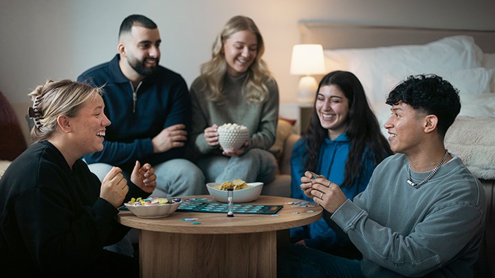 Fem glada studenter sitter runt ett bord. De spelar ett spel och fikar.