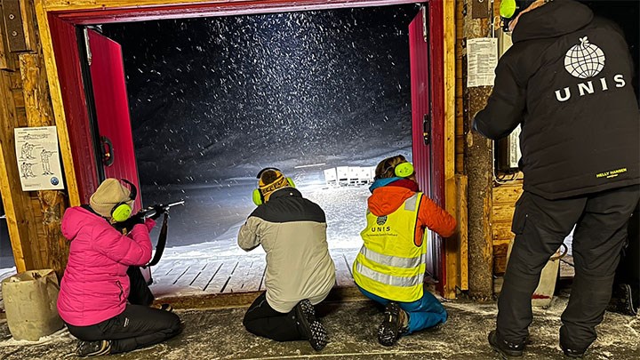 Three people on their knees indoors, being instructed to fire their rifles through a door into the night.