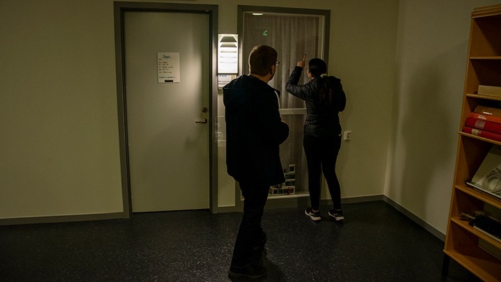Två personer kikar in genom ett fönster från en mörk korridor. En av dem pekar mot en glömd lampa.