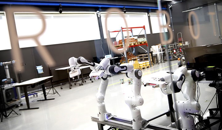 En bild tagen in genom fönstret på universitetets robotlabb. Man ser vita industrirobotar och en större lokal - på rutan står det "Robot".