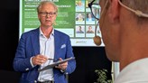 En man står med blå kavaj och ljus skjorta. Han håller en digital platta i handen. Med ryggen mot fotot står en annan man, snett bakifrån. Han har ett headset på huvudet. 