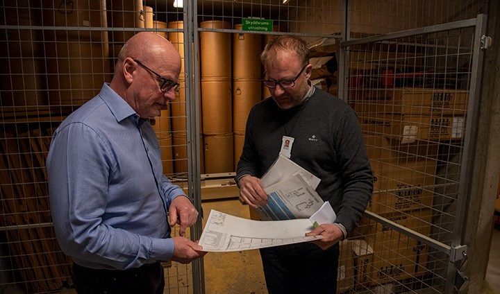 Skyddsrumsplaner på papper gås igenom av Owe Flodin, fastighetschef på Akademiska hus och Erik Nilsson, säkerhetschef på Örebro universitet. De står framför ett skyddsrumsförråd med bruna vattentunnor.