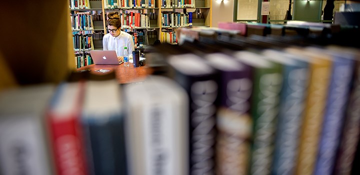 Photo of a person working with a laptop surrounded by bookshelves.