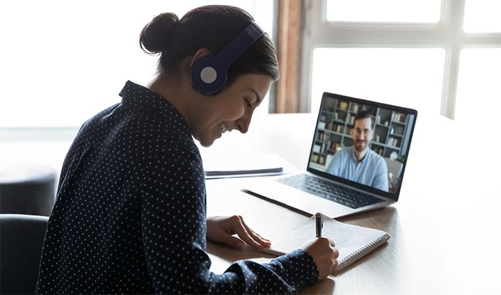 En kvinna sitter i digitalt möte framför en laptop och antecknar.