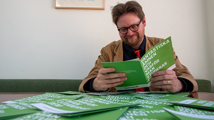 Peter Andersson läser broschyren "Fantastiska ämnen och varför man studerar dem". Framför honom  finns ett bord fyllt av samma broschyr.