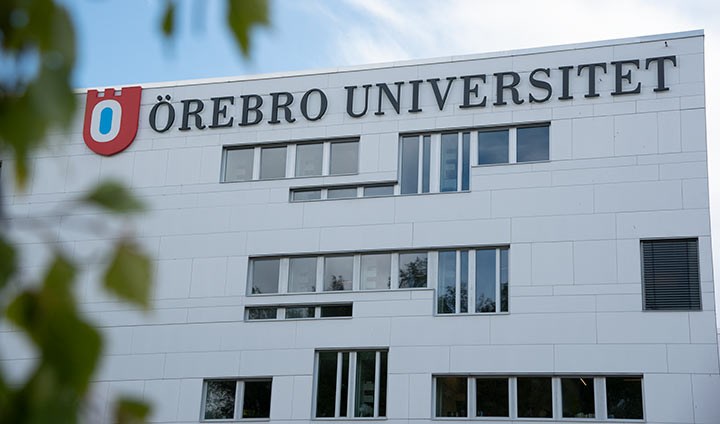 Vit byggnad med texten Örebro universitet. I förgrunden syns gröna löv.