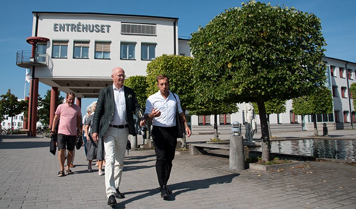 Johan Schnürer och Daniel Andersson går framför en vit byggnad med texten "Entréhuset". Till höger i bild syns grönskande träd klippta i kubform.