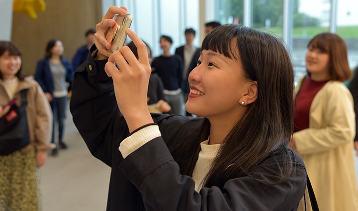 A female student smiles as she takes a picture with her mobile phone. In the background, there are other students from the Japanese group.