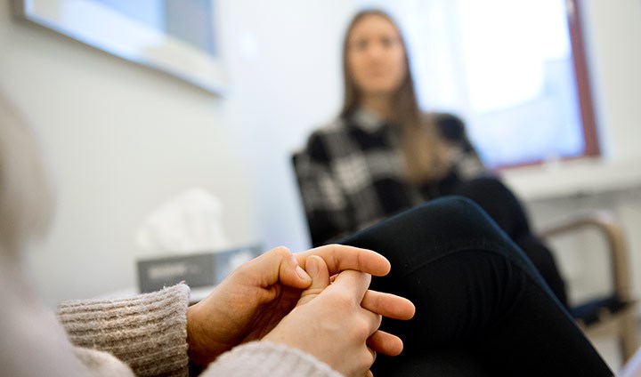 En bild från en psykologmottagning. I förgrunden syns ett par spända händer, i bakgrunden en suddig person i en stol och en låda med pappersservetter på ett bord.