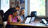 Foto på en lärare som står vid två sittande elever som har varsin bärbar dator.