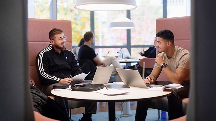 Två studenter sitter med datorer i biblioteket.