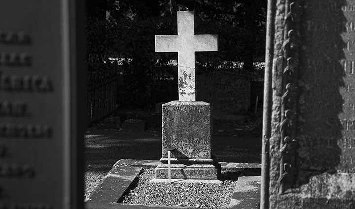 En svartvit bild på en gravsten i form av ett vitt kors. Vid sidan syns två svarta gravstenar.