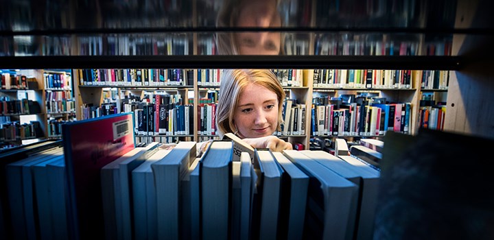 Foto på en person som står och letar bland böcker i en bokhylla.
