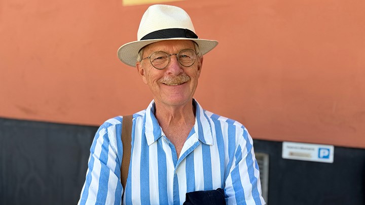 Erik Amnå i hatt och blåvitrandig skjorta. Han ler och står mot en rostfärgad och svart vägg. 
